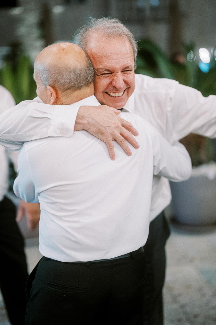 Two older men in white dress shirts warmly embrace each other with joyful expressions, capturing a moment of friendship and happiness at a formal gathering.