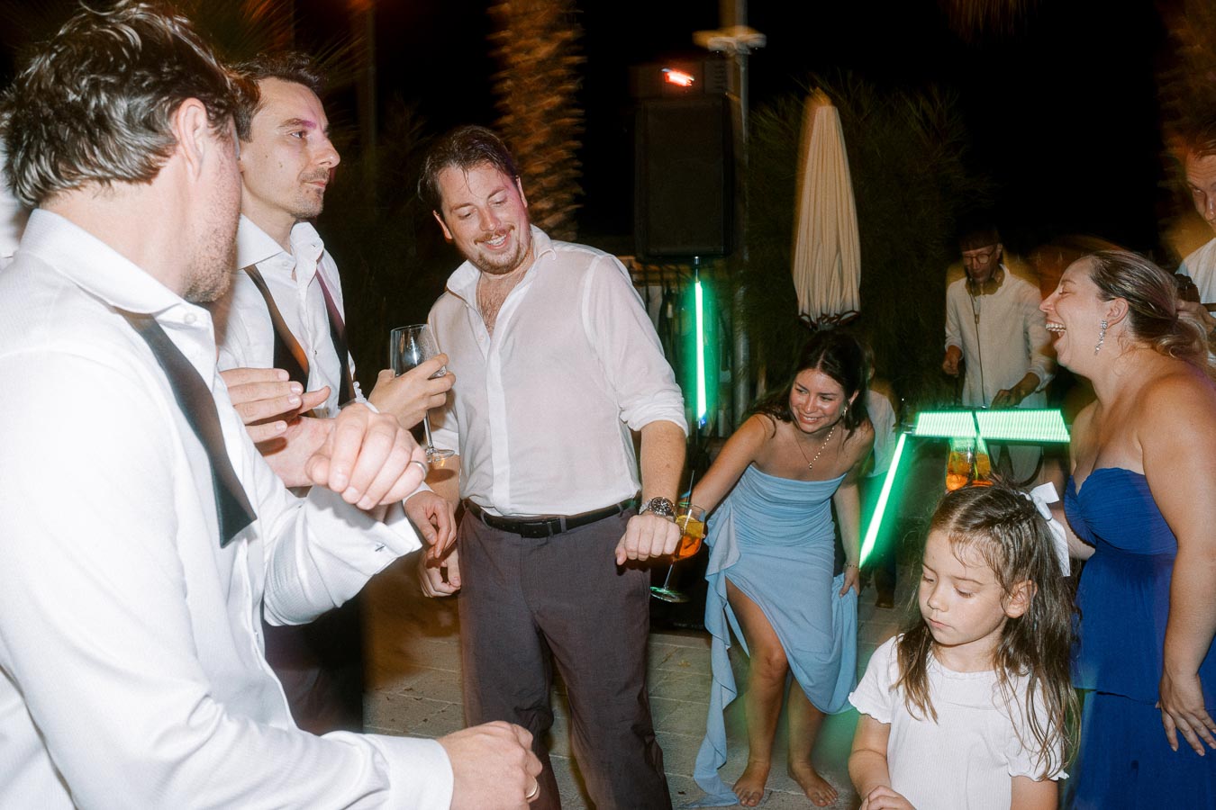 A group of people, including men and women, are joyfully dancing and socializing at an outdoor evening event. The scene features festive lighting, elegant attire, and a lively atmosphere. A young girl stands in the foreground, adding to the cheerful setting.