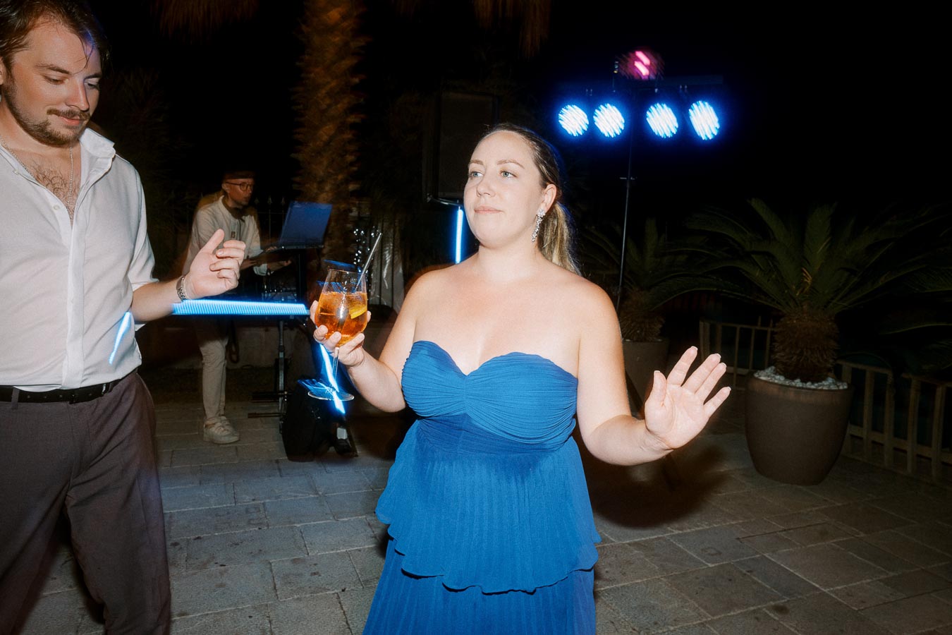 A woman in a blue strapless dress holding a cocktail at an outdoor evening party with colorful lights and a DJ in the background.