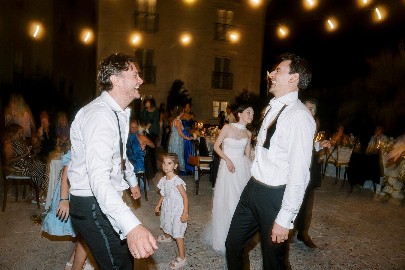 Outdoor evening wedding celebration with guests dancing energetically. Men in formal shirts and loosened ties enjoy the ambiance under string lights, while a woman in a white dress joins the festivities. Young girl in foreground, surrounded by people sitting and mingling in the background.