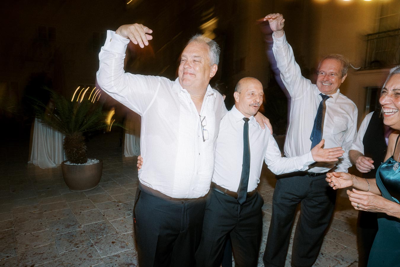 Group of people dancing and enjoying a celebration outdoors at night, with blurred motion to capture lively movement, wearing formal attire and smiling.