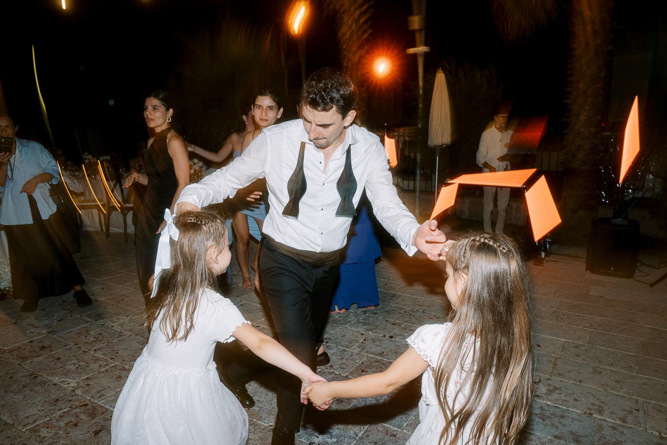 A man in formal attire dances with two young girls in white dresses at an outdoor evening event, surrounded by other guests and illuminated by warm lights.