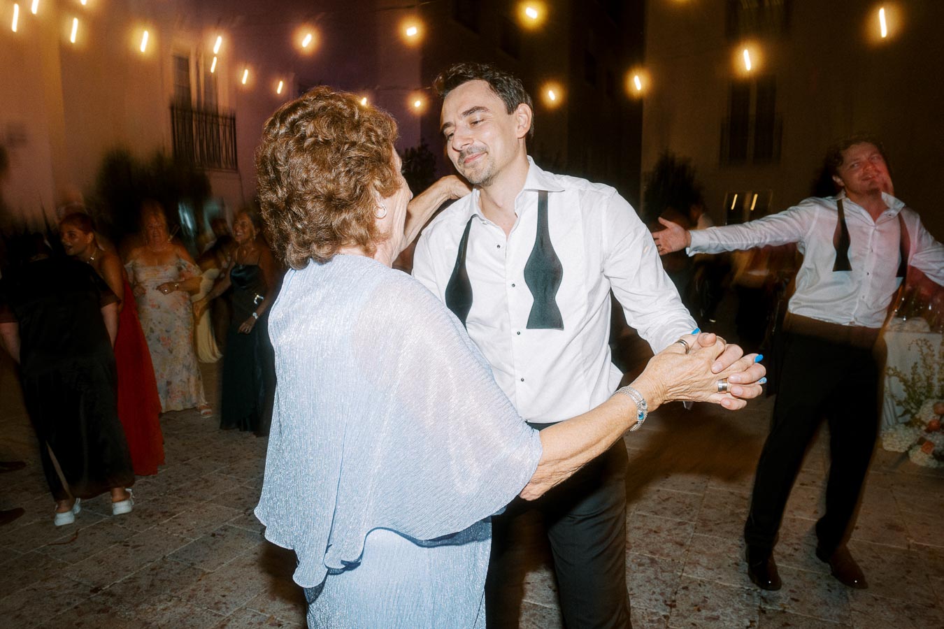 Elegant evening dance scene with a man and older woman dancing under softly lit string lights, surrounded by guests in formal attire at an outdoor event.