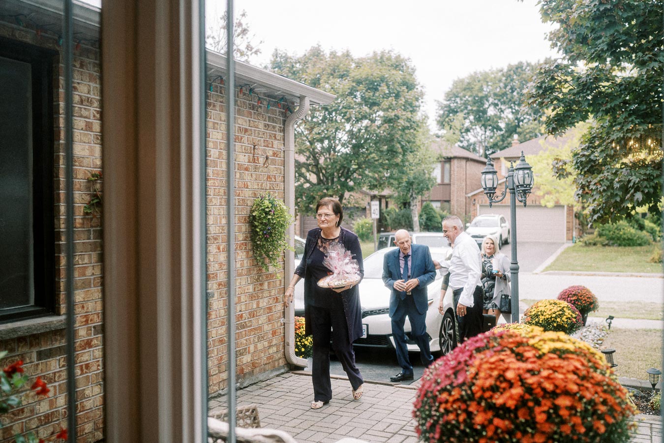 A group of well-dressed people arriving at a house entrance, one woman carrying a wrapped gift basket, with colorful potted