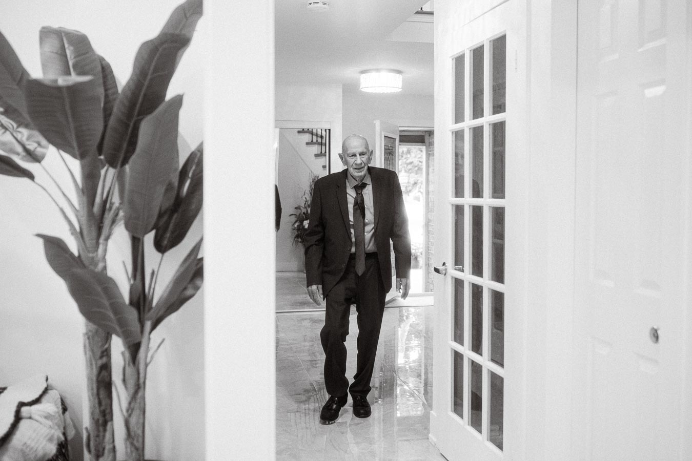A black and white photo of a man in a suit walking through a modern home interior with a large houseplant in the foreground