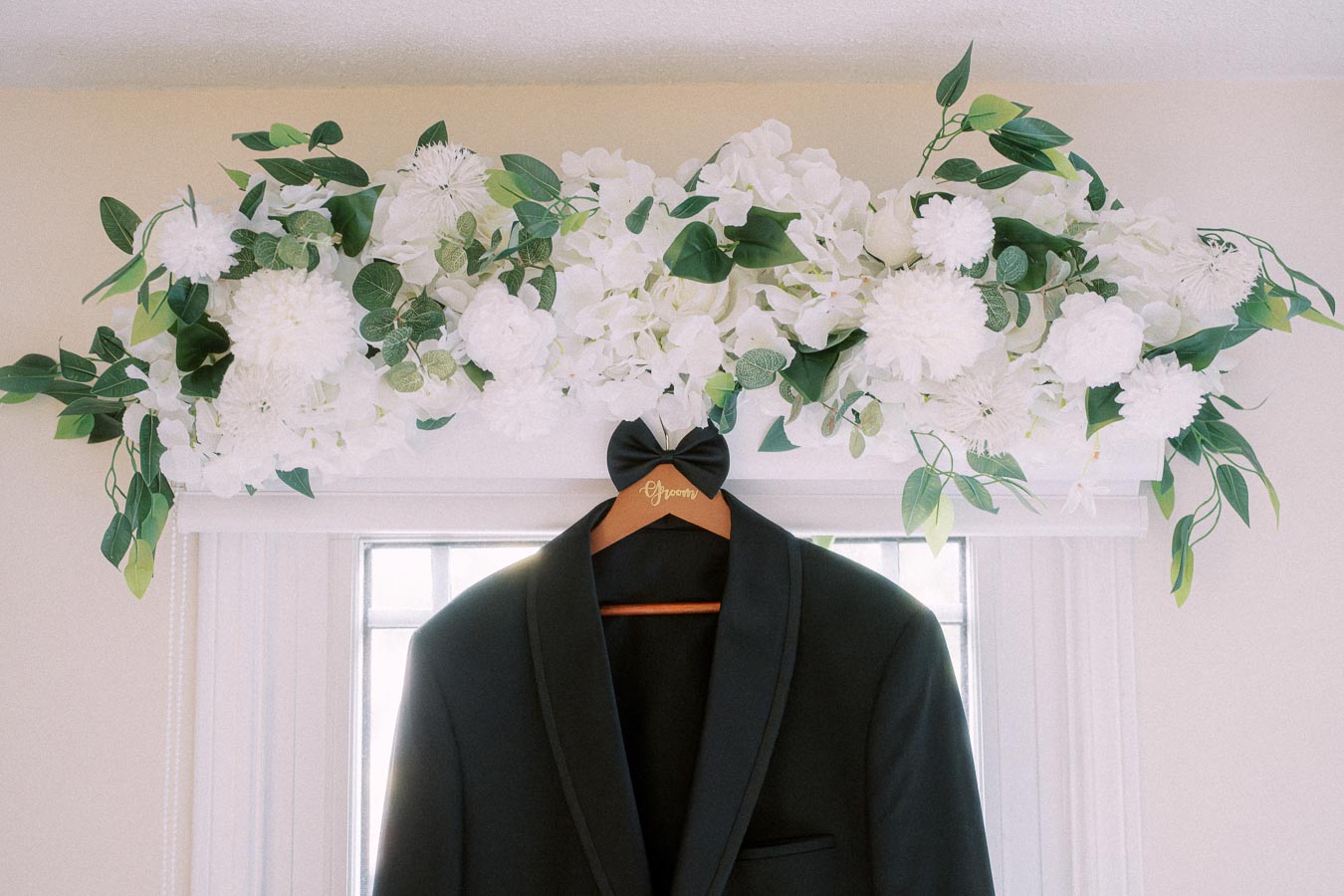 Elegant black tuxedo hanging on a wooden hanger labeled Groom beneath a floral arrangement of white flowers and green