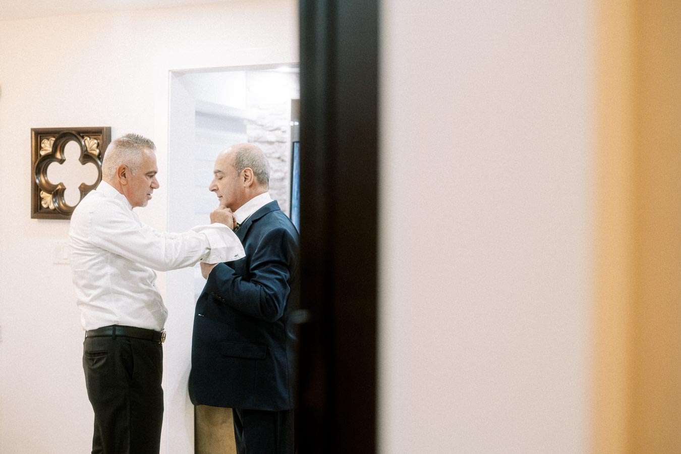 Two men dressing formally in a room, one adjusting the other's tie, symbolizing friendship and preparation for an event.
