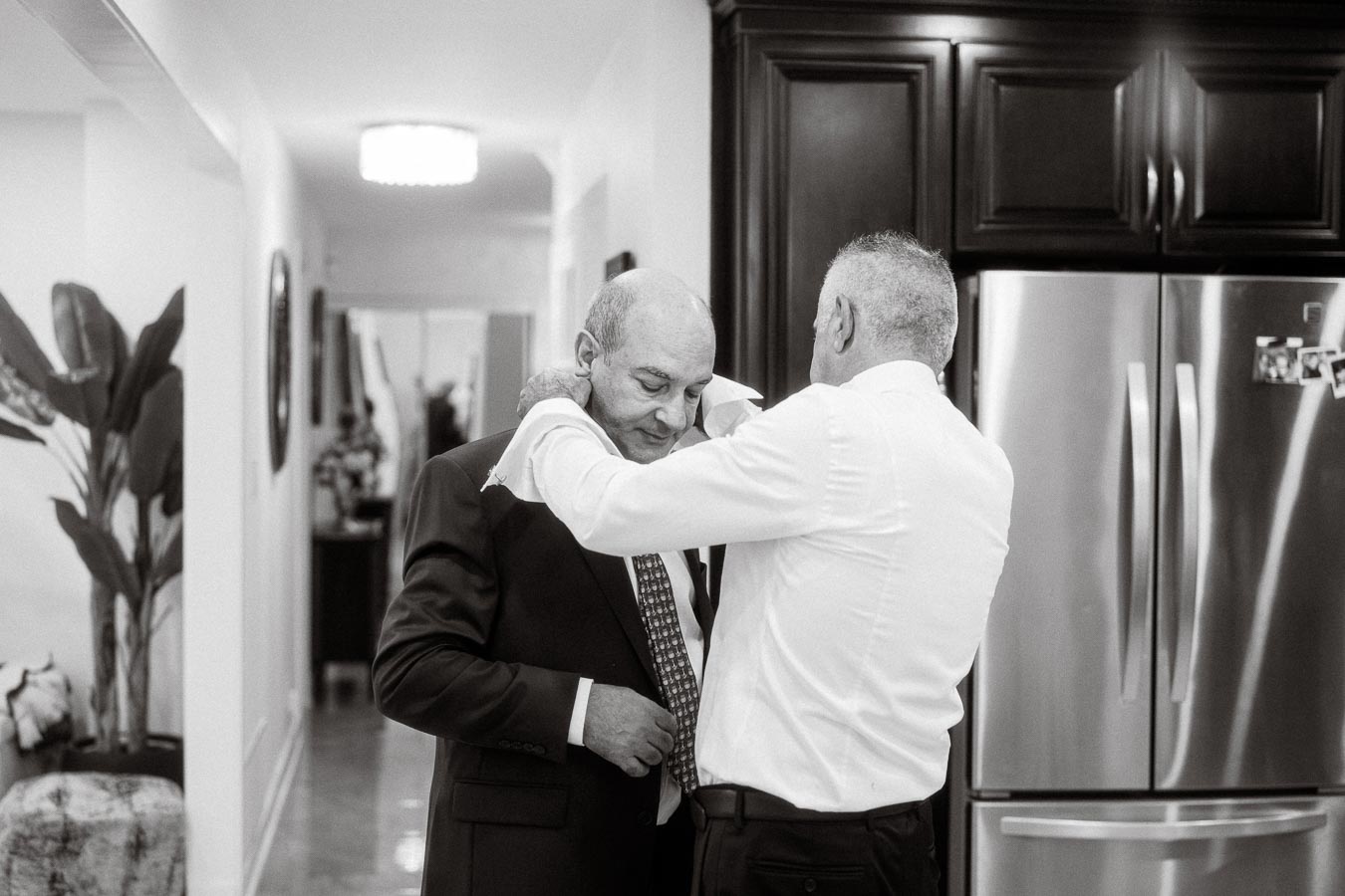Two men in a modern kitchen, one helping the other adjust his suit jacket, depicting a moment of preparation and friendship.