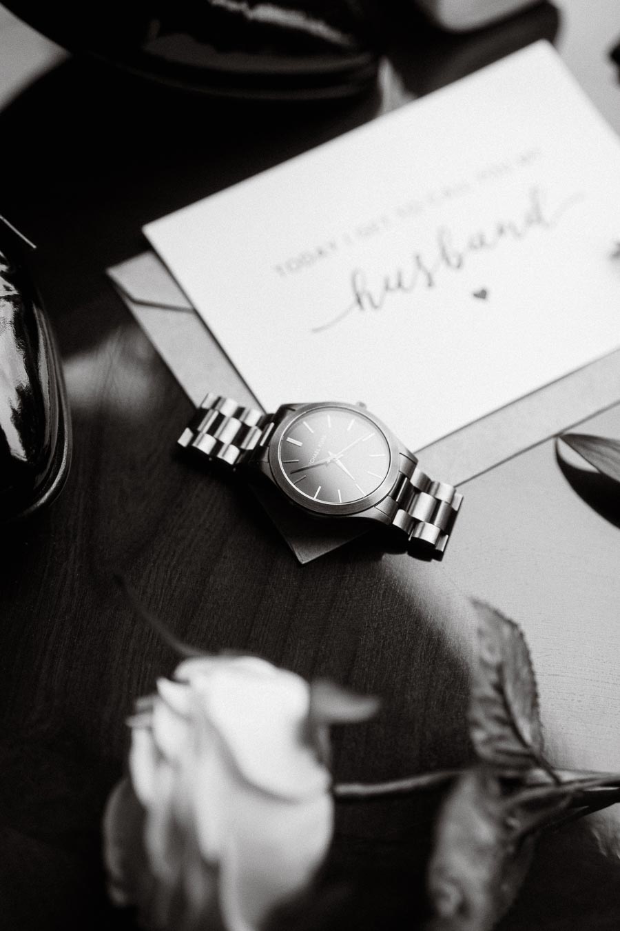 Black and white image of a luxury wristwatch placed on a table, alongside a pair of polished dress shoes and an elegant card