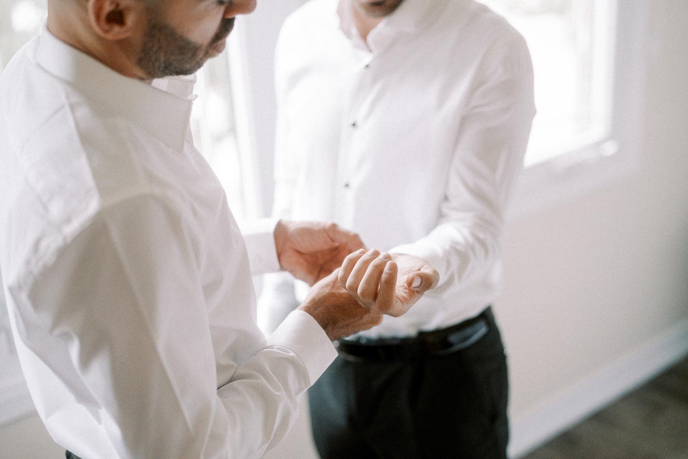 Two men dressed in white shirts and black trousers, engaging in a close conversation indoors near a window, with one man