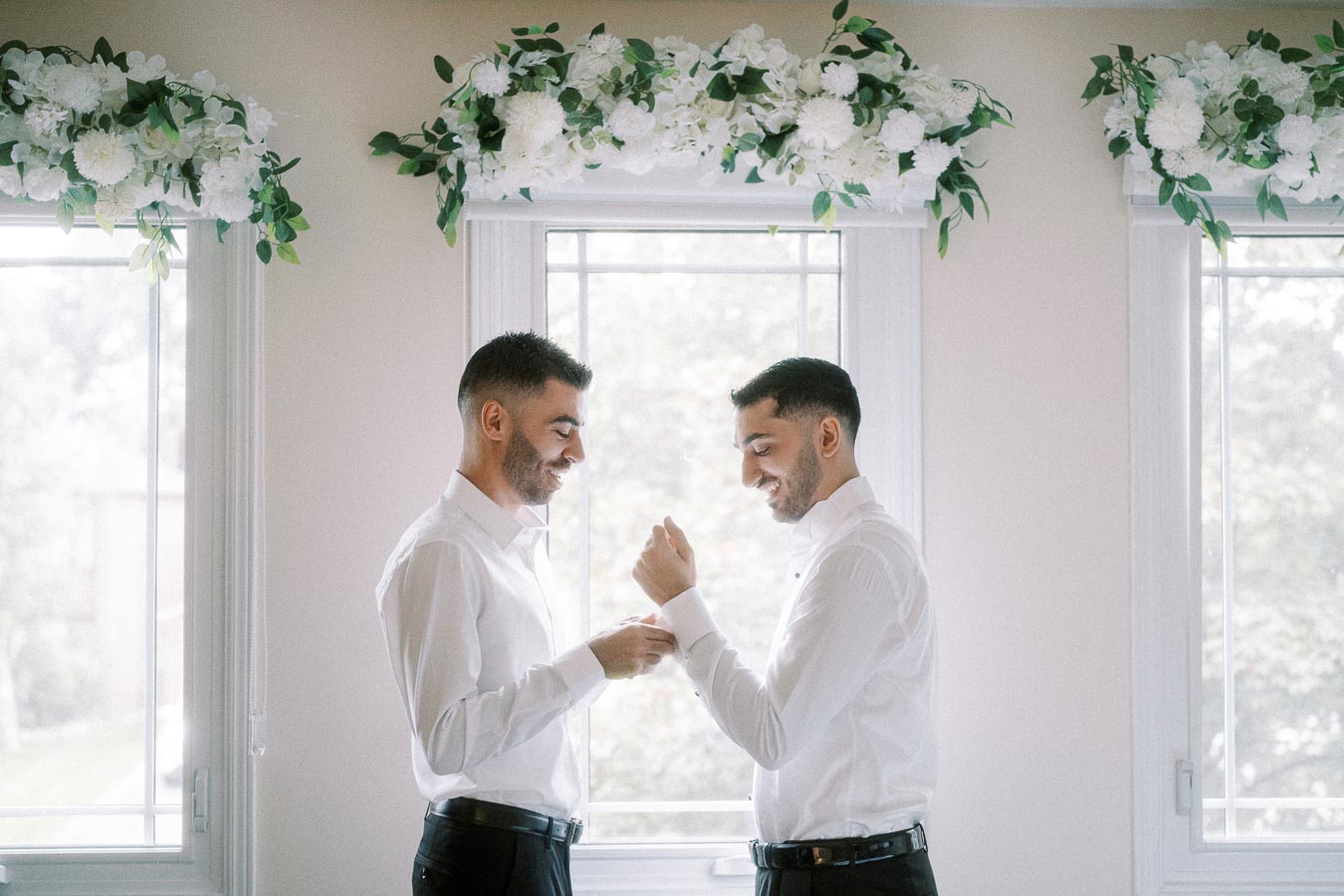 Two men in white shirts smiling and adjusting each other's cuffs in a bright room with floral decorations above the windows.