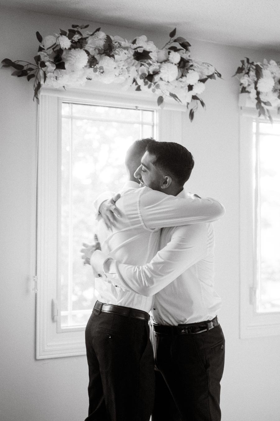 Two people embracing warmly near a sunlit window with floral decorations, wearing formal attire.