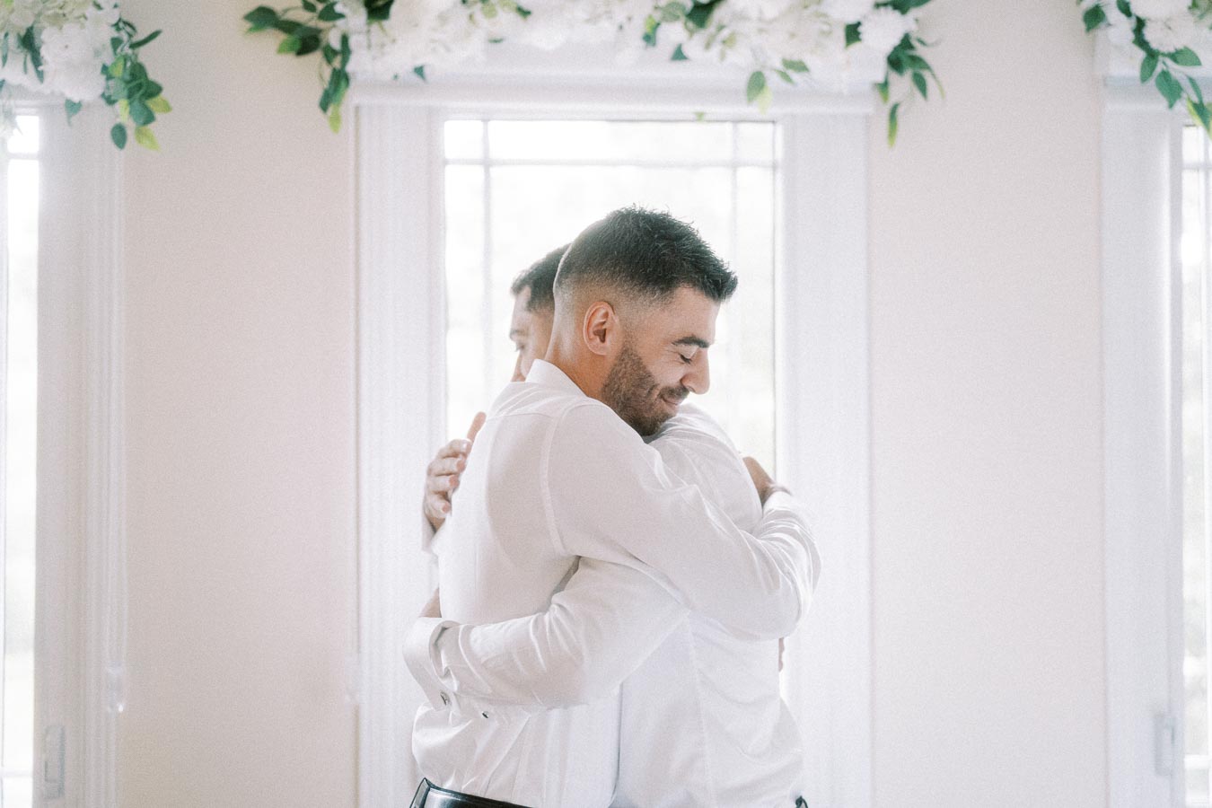 Two men in white shirts embracing warmly in a sunlit room with floral decorations above windows.