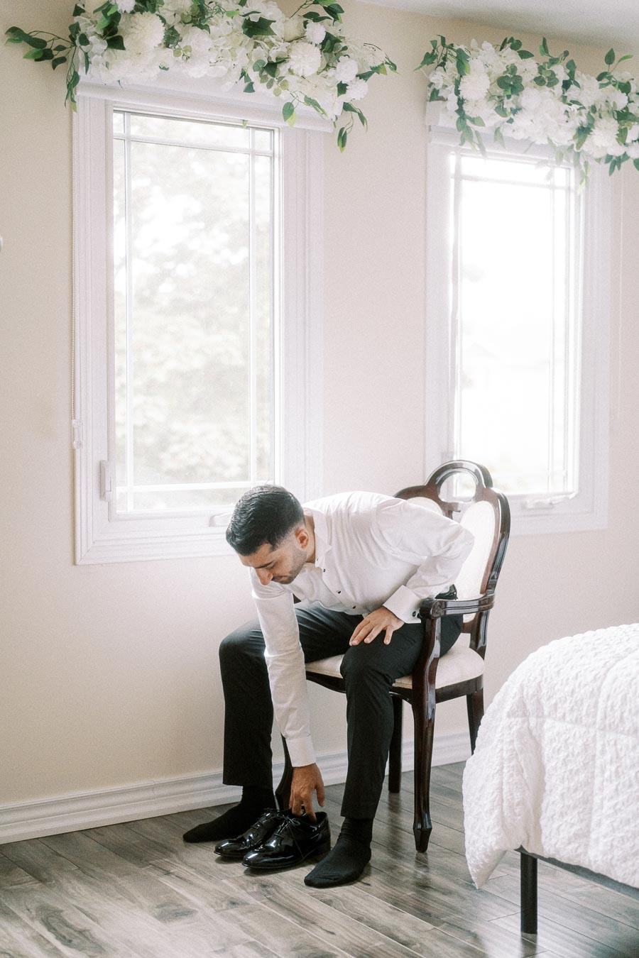 Groom in white shirt sitting on a chair, putting on black dress shoes, preparing for a wedding in a bright room with floral