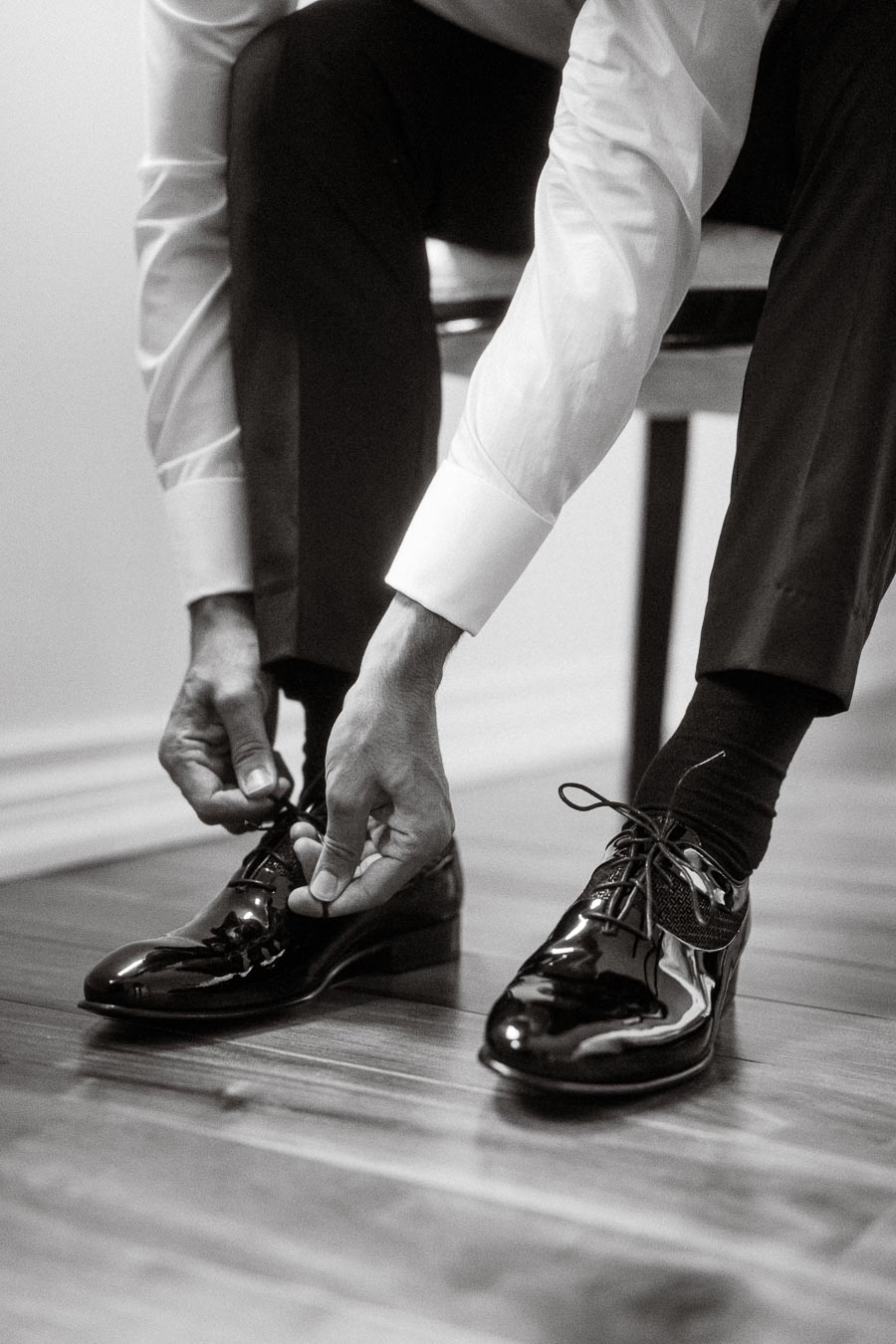Man tying shoelaces on polished black dress shoes, dressed in formal attire, sitting on a chair.