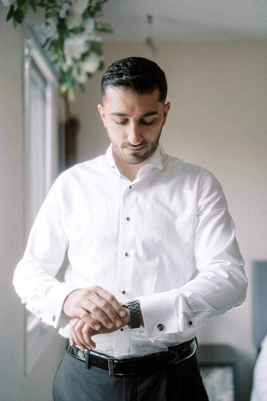 A man in a white dress shirt adjusting his watch, standing in a softly lit room with green foliage in the background,