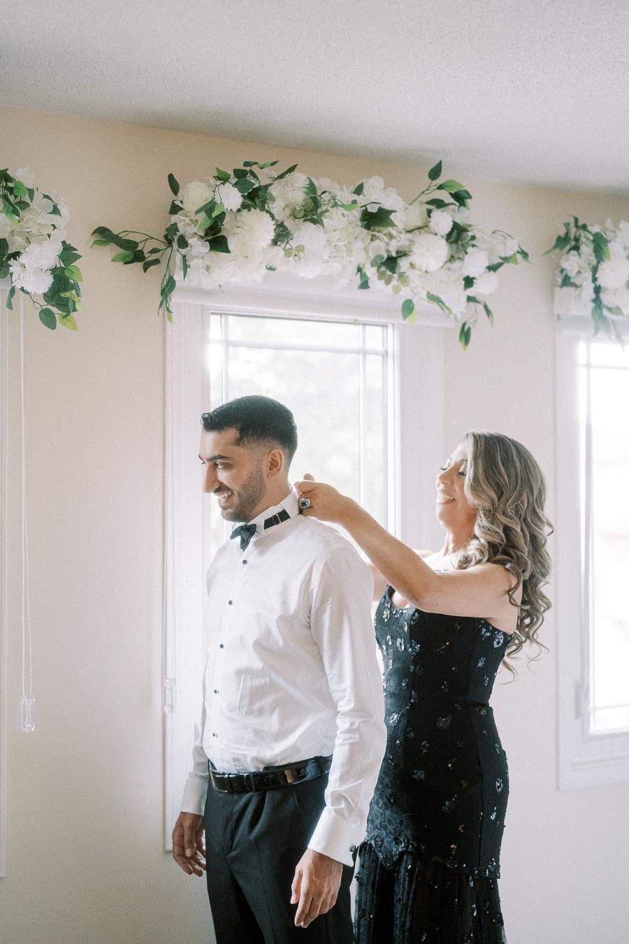 A smiling woman in a black dress adjusts a man's bow tie in a bright room decorated with white floral garlands, capturing a