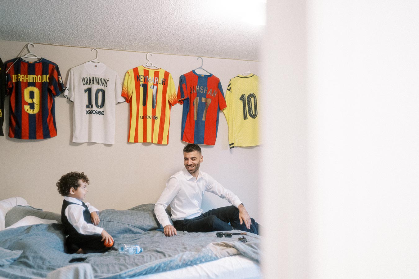 Father and son sitting on a bed in a room with soccer jerseys displayed on the wall, showcasing passion for football.