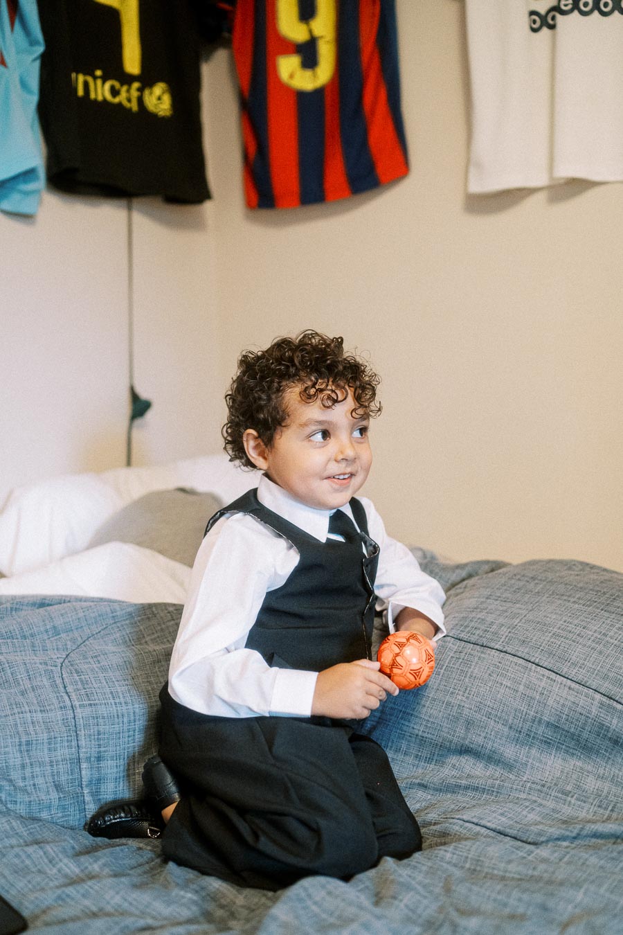A young boy with curly hair, dressed in a formal suit, sits on a bed holding an orange ball, with football jerseys hanging
