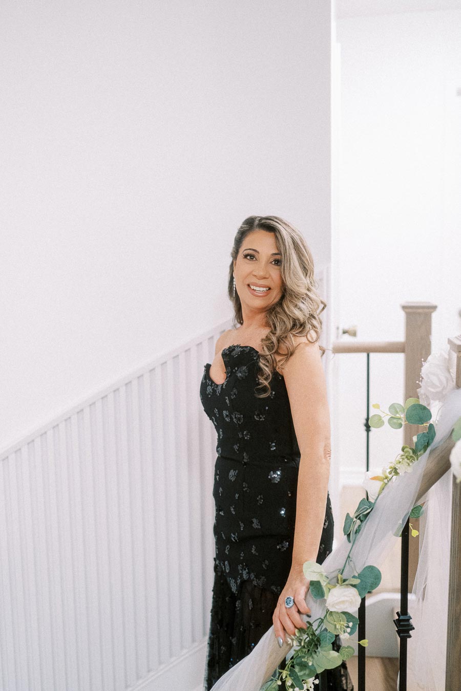 Smiling woman in elegant black dress standing on staircase decorated with greenery and white flowers.