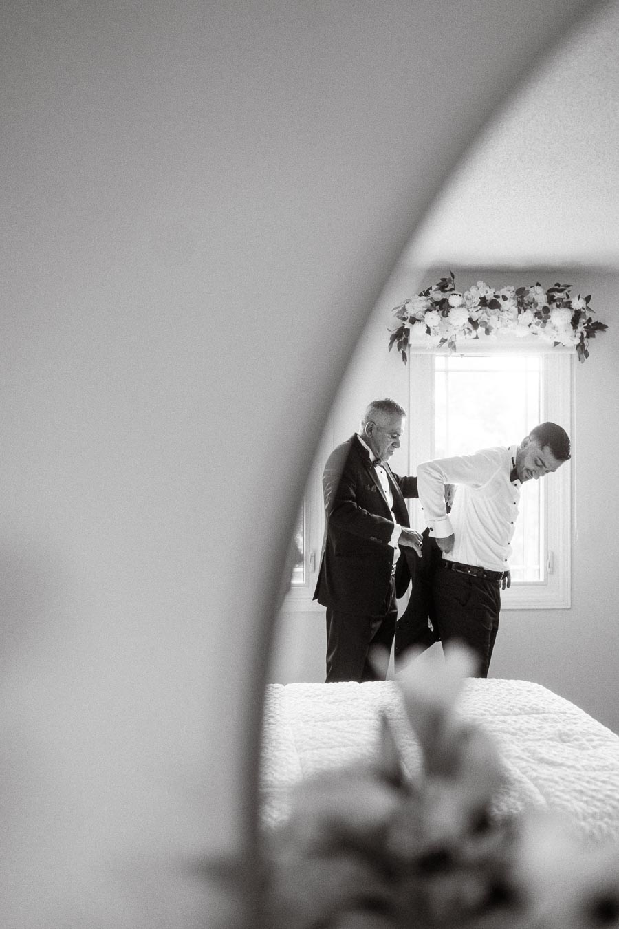 A black-and-white photo of two men dressing in formal attire next to a window adorned with floral decorations, captured