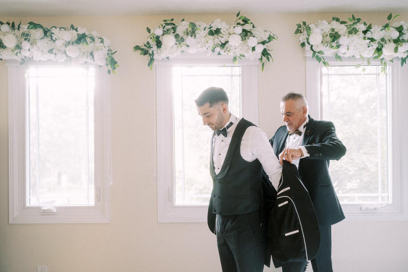A groom getting dressed for a wedding with the help of an older man in a room decorated with white floral arrangements above