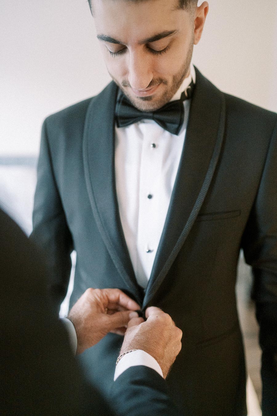 A groom wearing a black tuxedo and bow tie has his jacket buttoned by another person, preparing for a wedding ceremony.