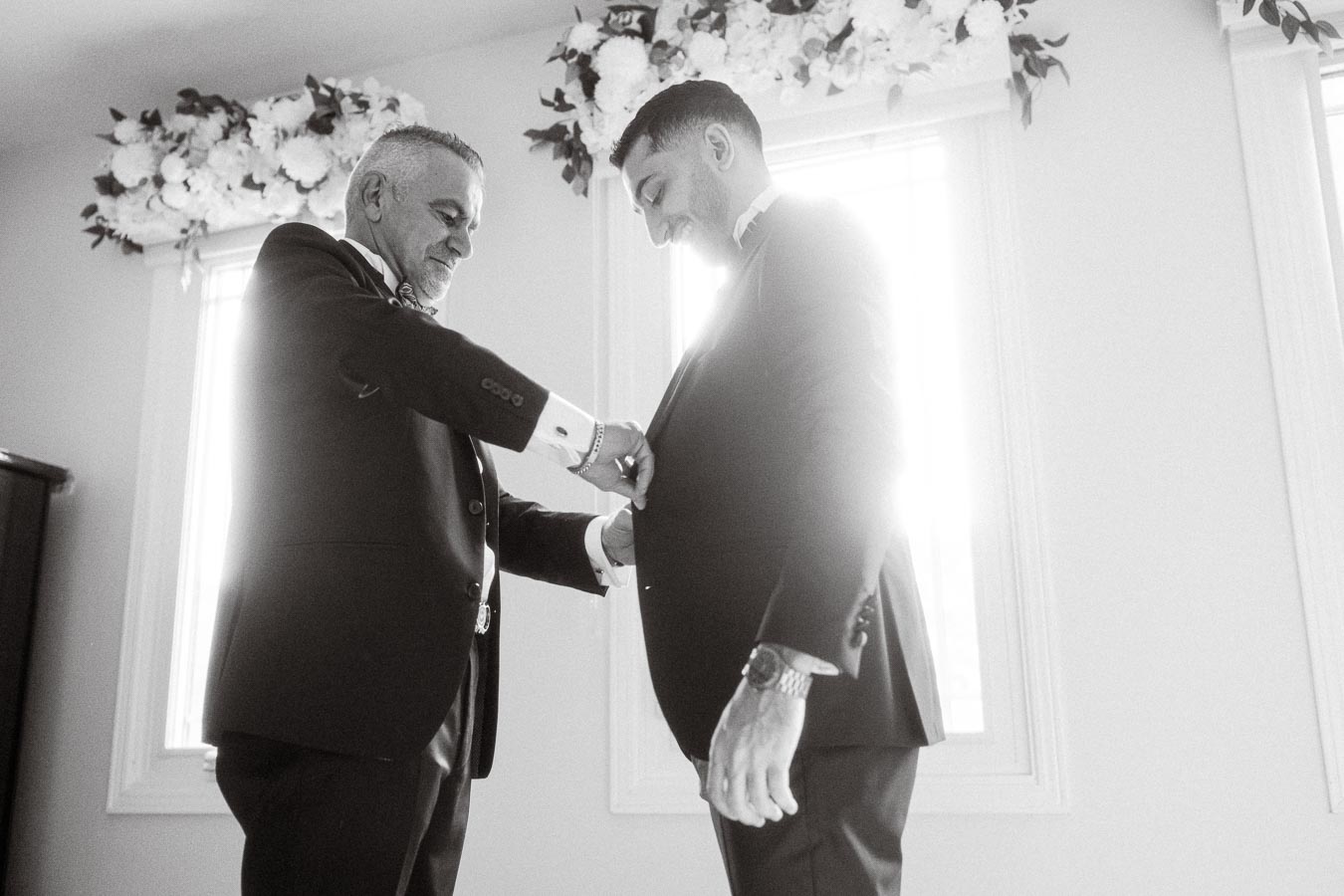 Two men in formal attire preparing for a wedding ceremony in a bright room with floral decorations, emphasizing the bond and