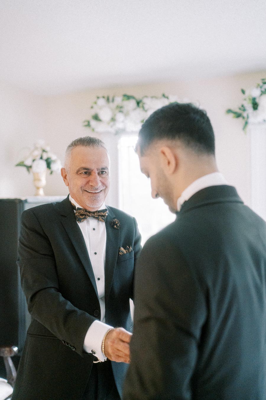 A joyful older man in a suit smiles while greeting a younger man also dressed in formal attire, in a beautifully decorated