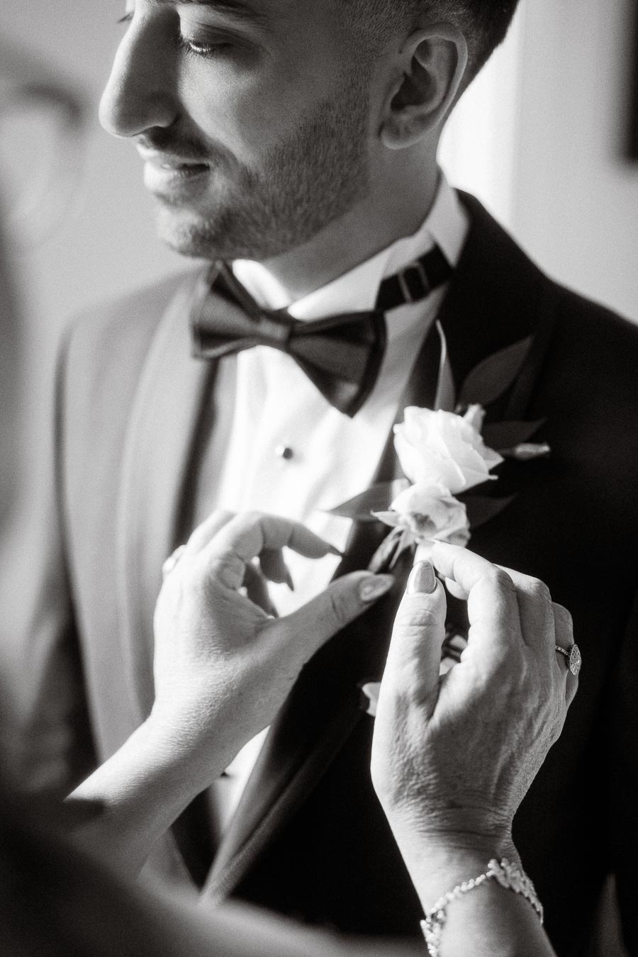 A groom in a suit and bow tie smiles while a helper pins a white boutonniere on his lapel, highlighting a special moment