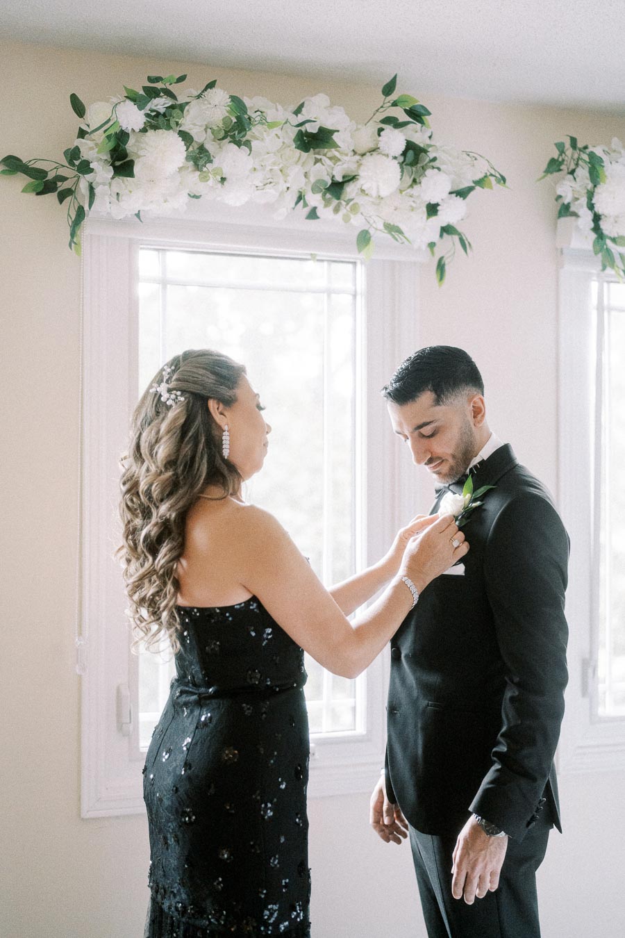 A woman in a black dress adjusts the flower on a man's tuxedo inside a room decorated with white floral arrangements.