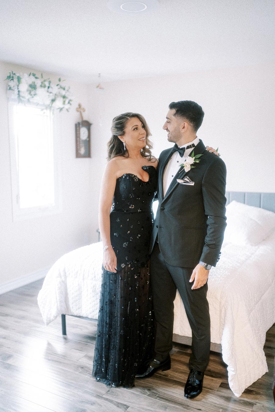 A couple in formal attire standing in a bright bedroom, with the woman wearing a black gown and the man in a black tuxedo,