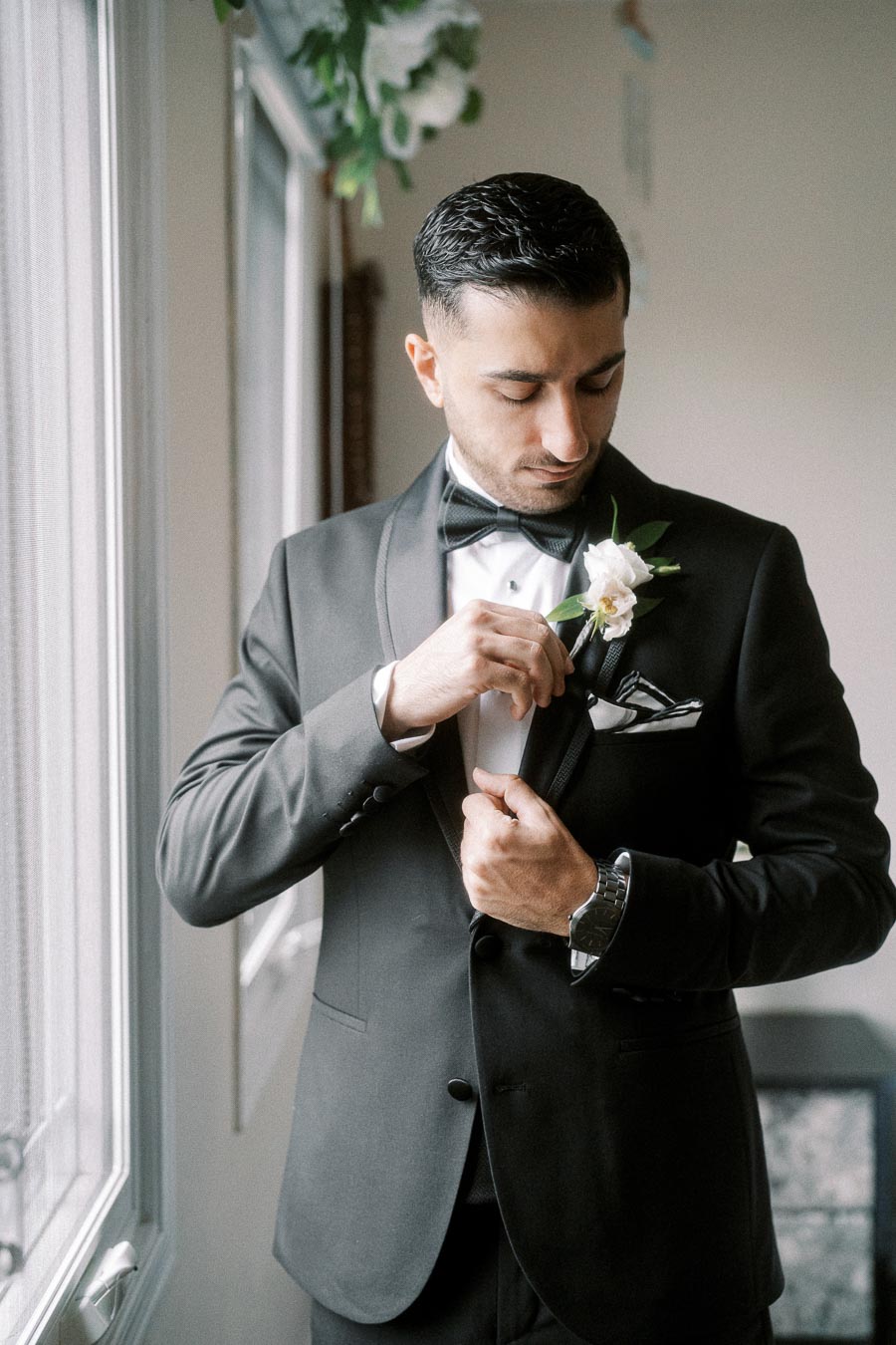 Elegant groom adjusting his black tuxedo jacket button before a wedding ceremony, with a white boutonniere and bow tie,
