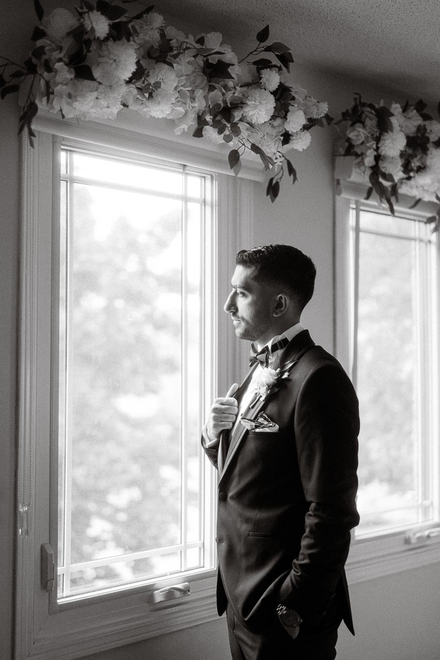 Black and white portrait of a groom in a suit standing by a window, adjusting his boutonniere, with floral decorations above