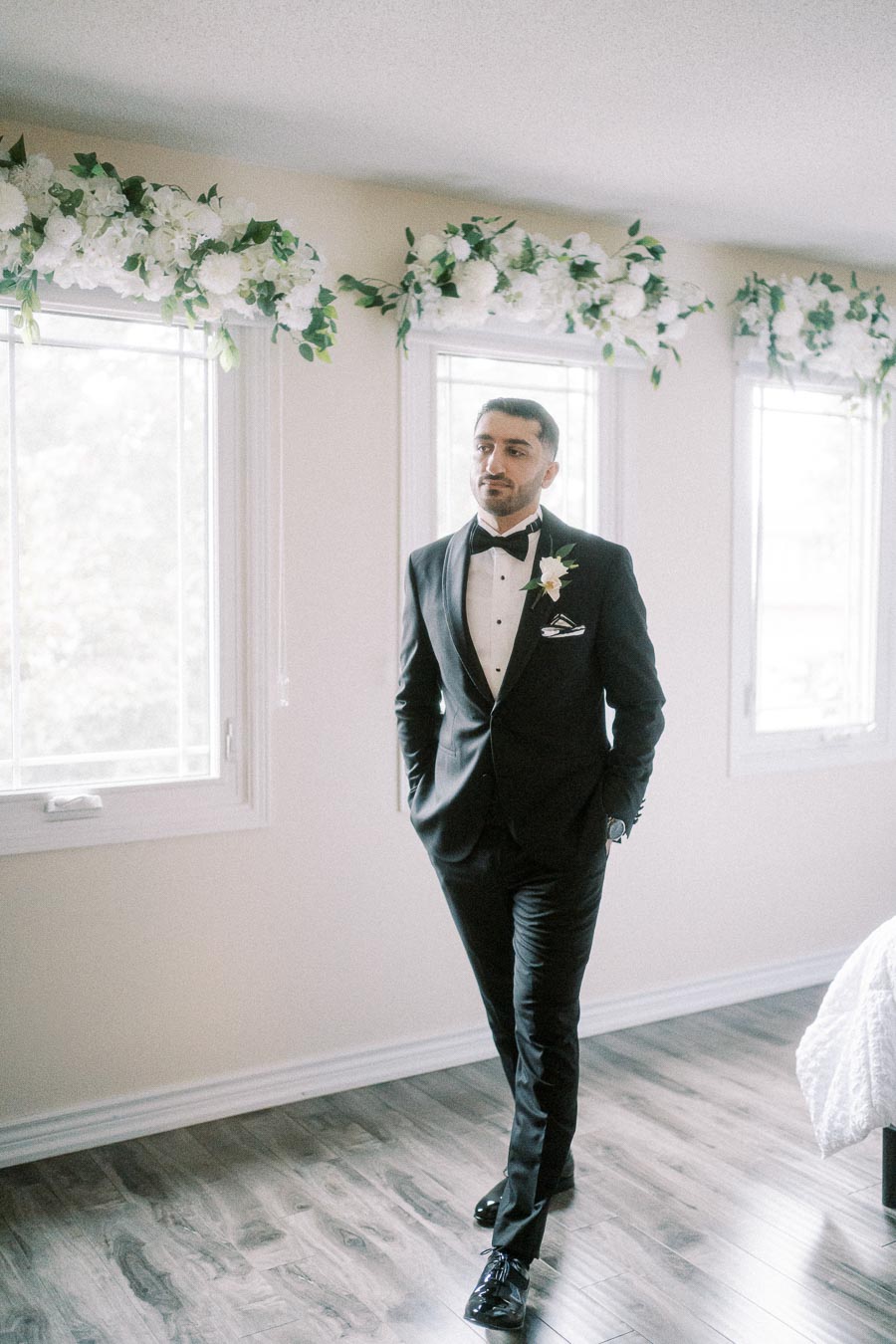 A groom in a black tuxedo with a white boutonniere stands confidently in a well-lit room with floral decorations above large