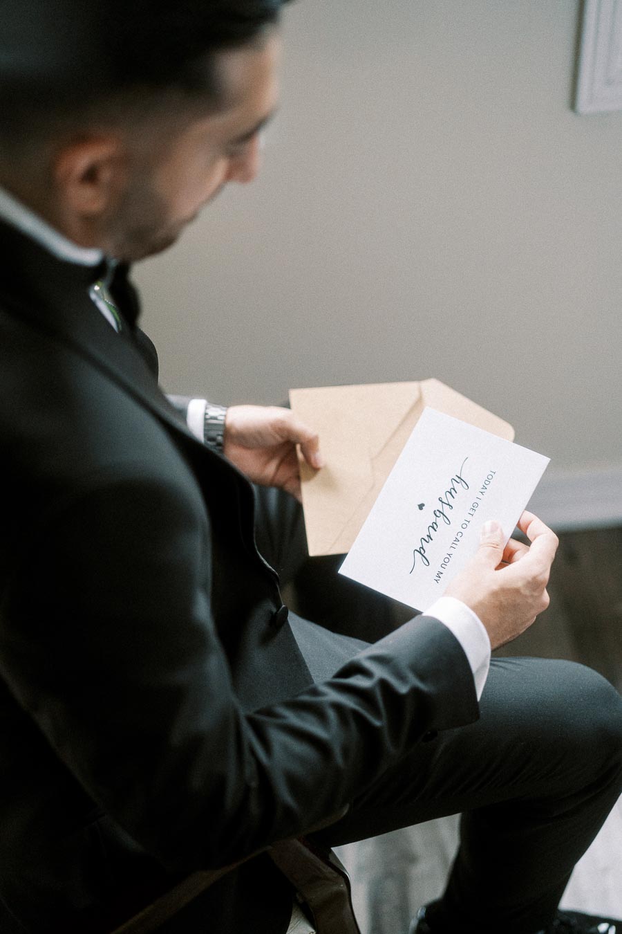 A man in a suit reads a wedding invitation with Today I get to call you my husband in elegant script.