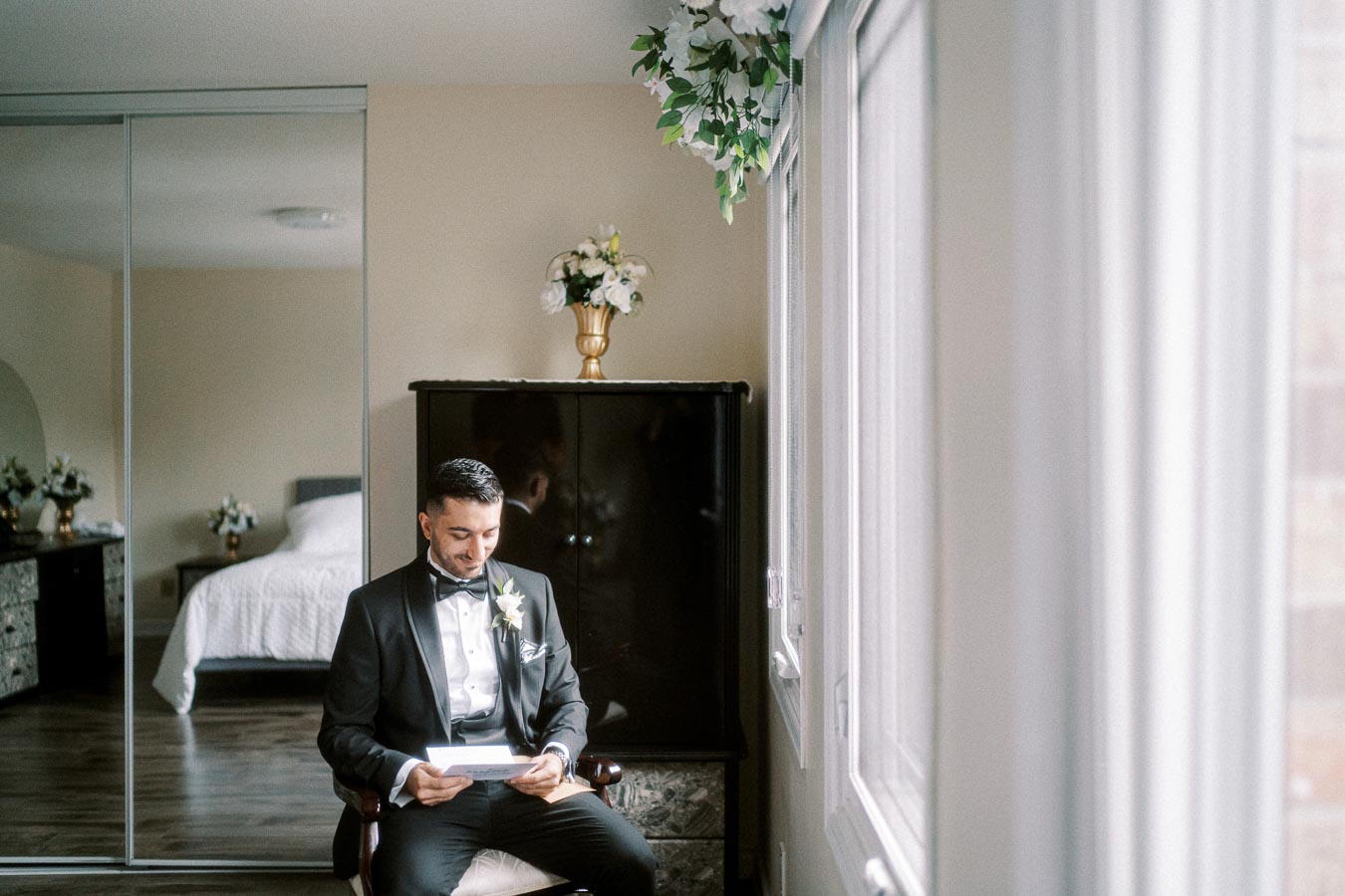 Groom sitting and smiling while reading a letter in a bright room, wearing a black tuxedo with a white boutonniere,