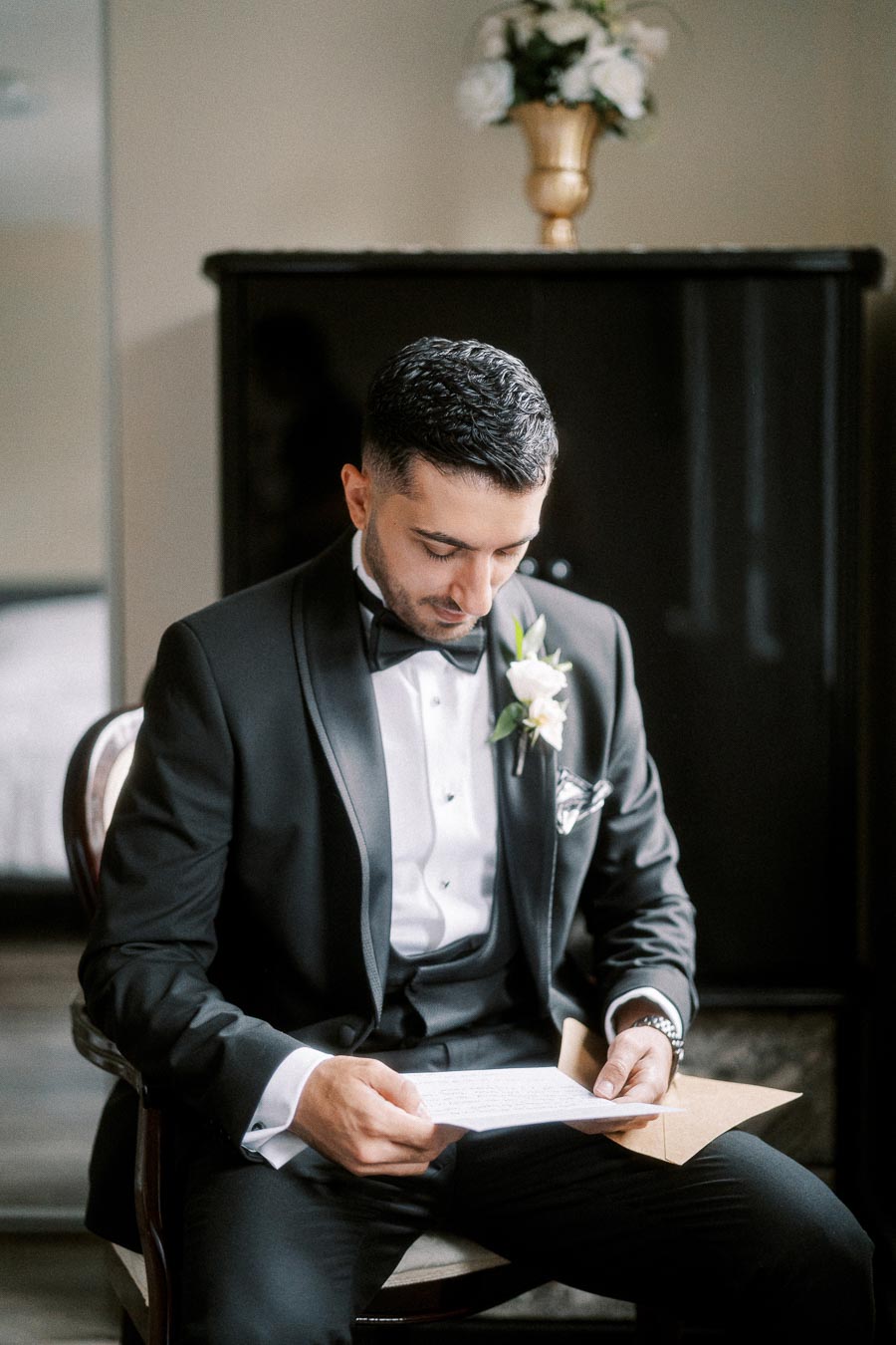 Groom sitting in a stylish black tuxedo with a bow tie, holding a letter and looking thoughtful on his wedding day, with a