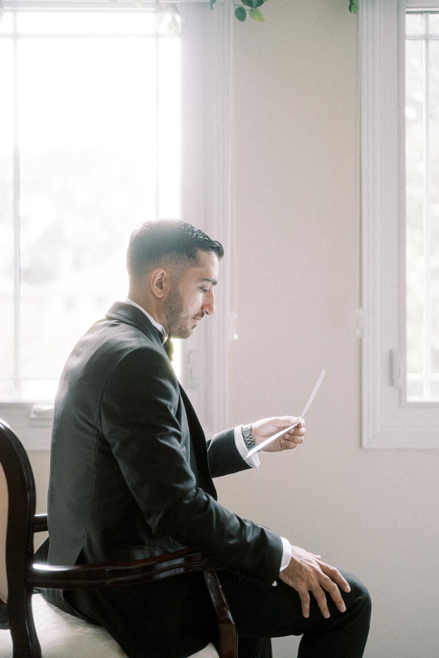 A groom in a suit sitting on a chair and reading a letter in a softly lit room with large windows.