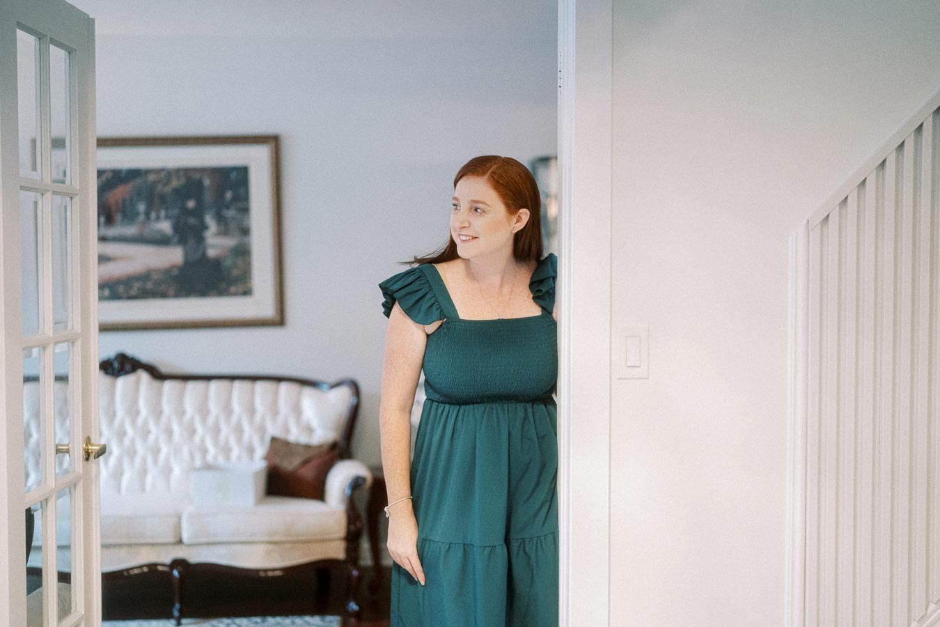 Woman with red hair smiling while standing in a doorway, wearing a green dress in a bright, elegantly decorated living room