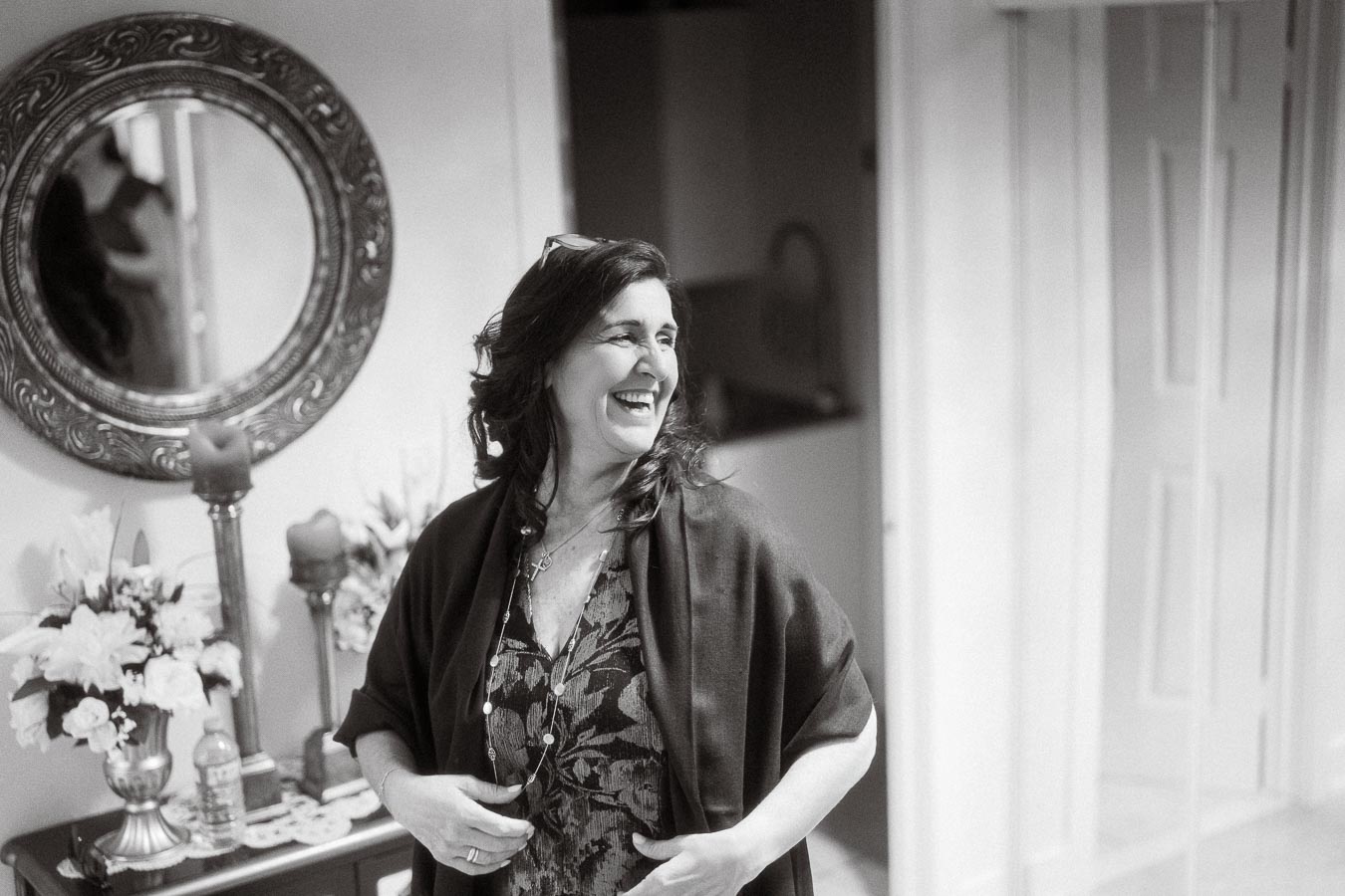 Black and white photo of a woman smiling in a beautifully decorated room with a decorative mirror, floral arrangement, and