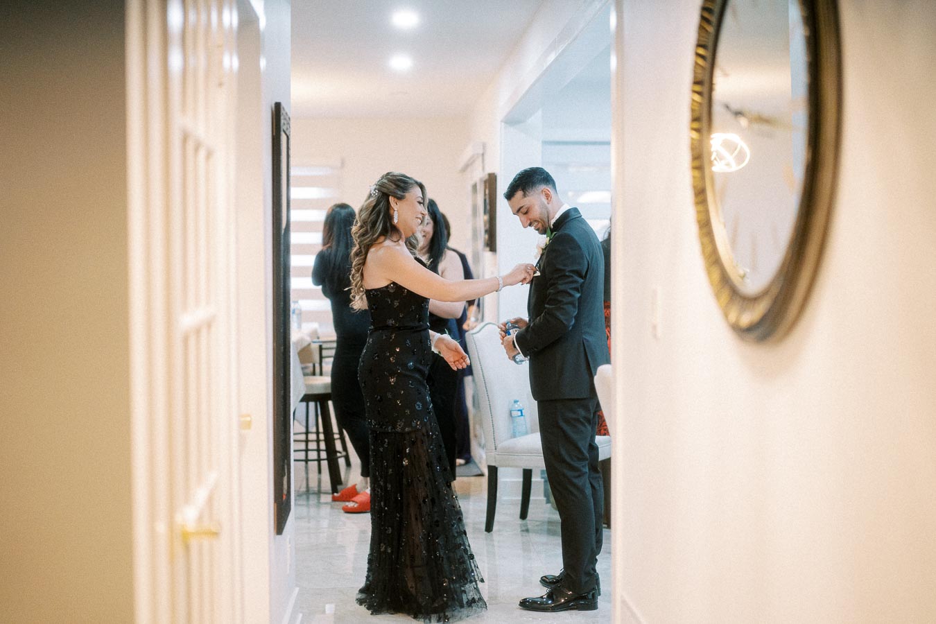 Couple in formal attire preparing for an event in a stylishly decorated hallway, with a woman adjusting a man’s boutonniere