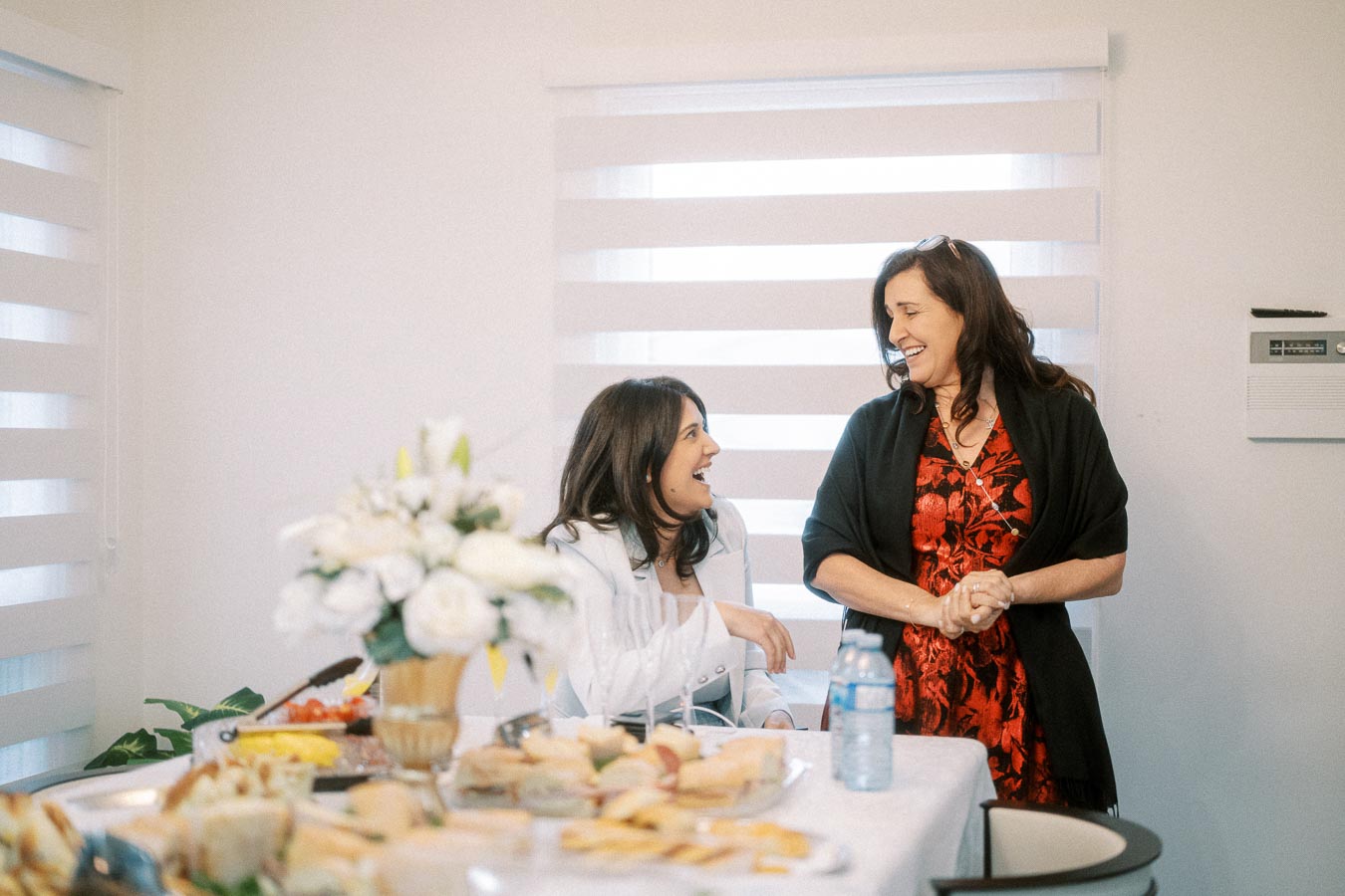 Two women smiling and conversing at a dining table adorned with a variety of sandwiches and a floral arrangement, in a