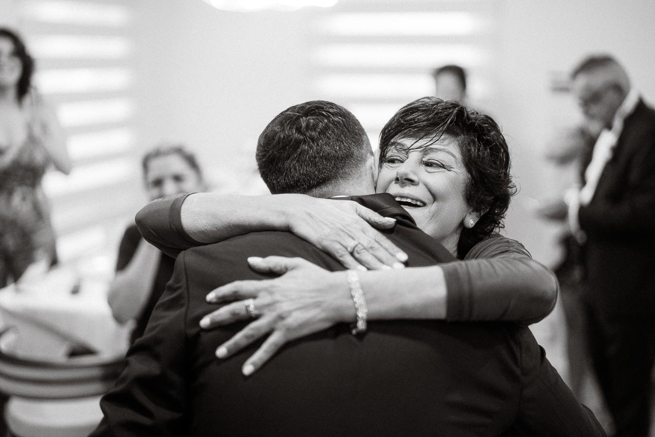 Black and white photo of a joyful woman embracing a man at a social gathering, with other people in the background,