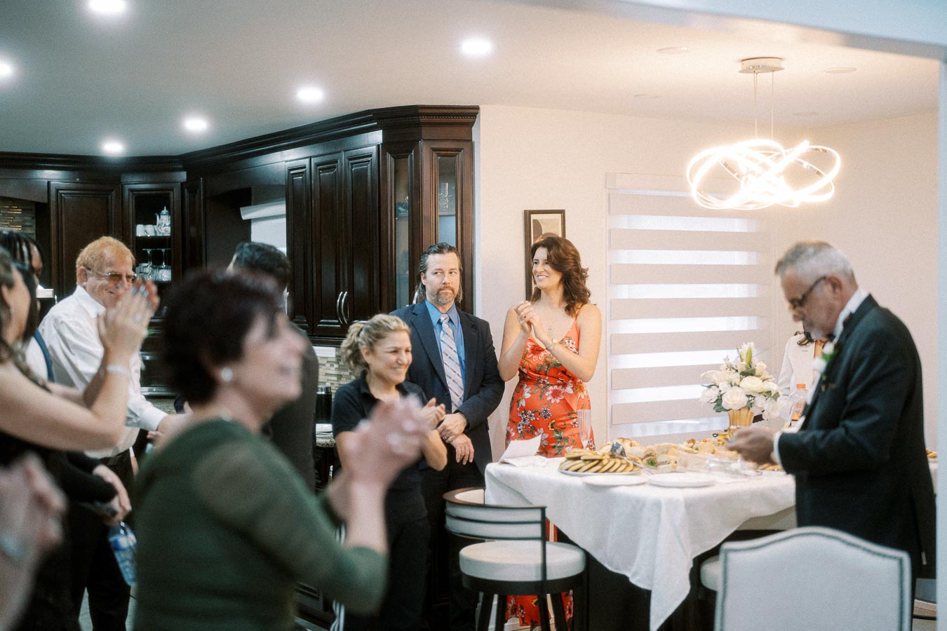 A group of people gathering in a modern kitchen, clapping and smiling, with one person in a floral dress near a table set