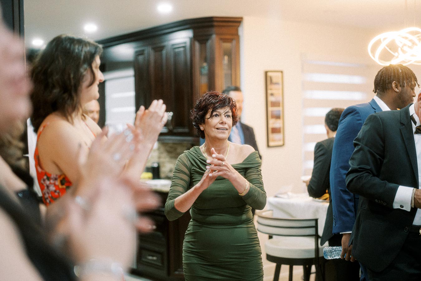 A group of elegantly dressed people celebrating indoors, focused on a woman in a green dress clapping and smiling,