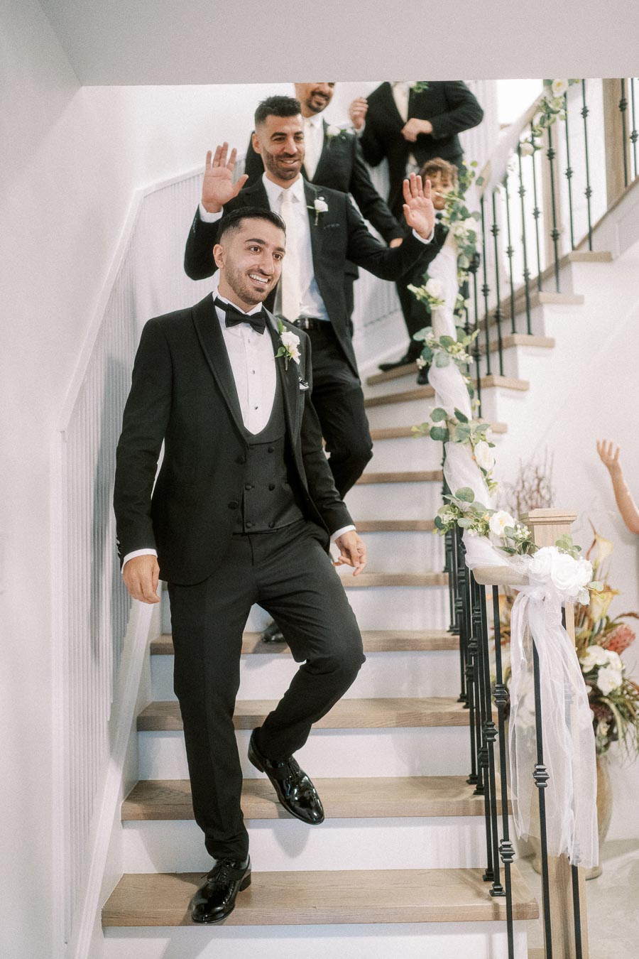 Groomsmen in elegant black tuxedos descend a beautifully decorated staircase with floral arrangements, exuding joy and
