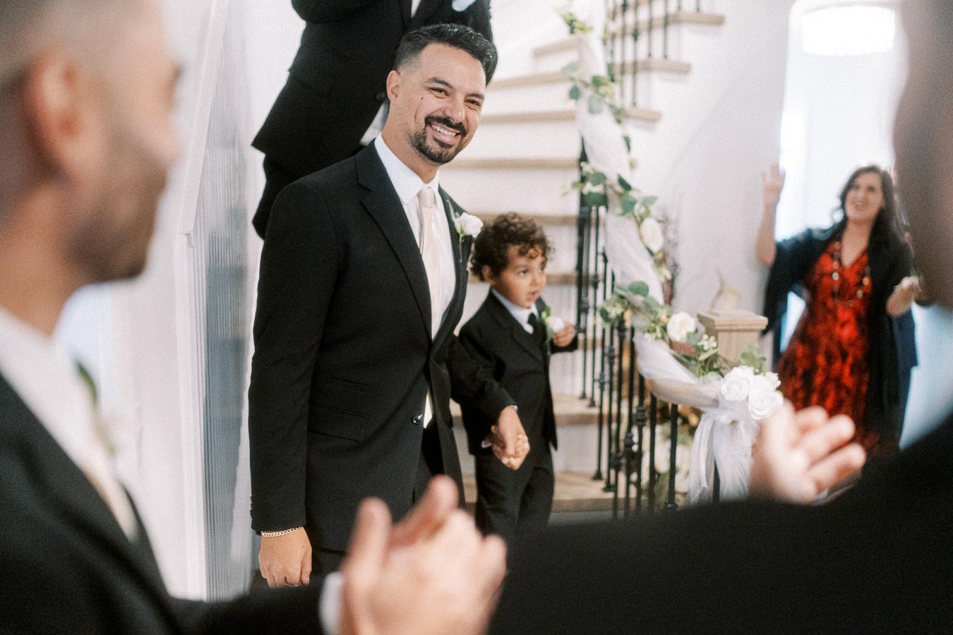 A smiling man in a suit holding the hand of a young boy, also in a suit, as they walk down a flower-decorated staircase