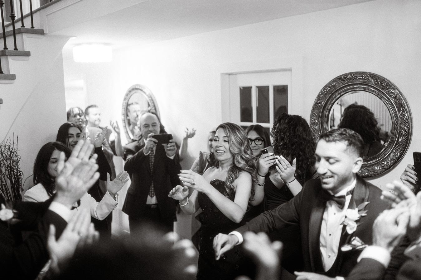 A joyful celebration with elegantly dressed guests clapping and photographing in a warmly lit room, featuring a woman in a