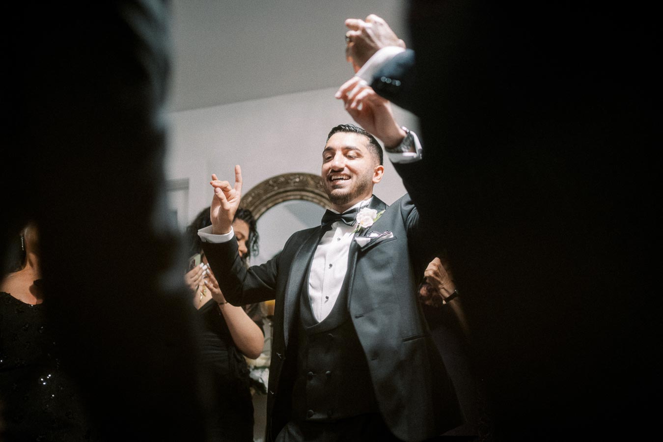 Smiling man in a tuxedo dancing at a formal event, surrounded by other guests, with a mirror in the background.